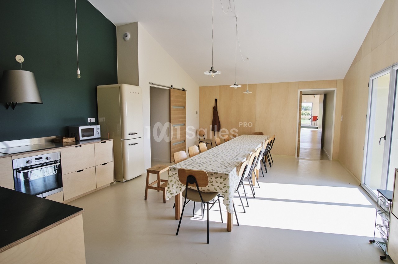 Cuisine moderne avec table longue, chaises en bois, électroménagers et lumière naturelle entrant par une baie vitrée.