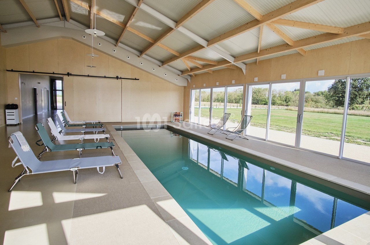 Piscine intérieure avec chaises longues, grandes baies vitrées donnant sur un jardin verdoyant.