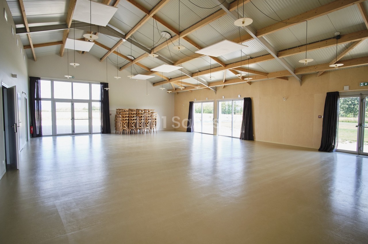 Grande salle lumineuse avec un sol beige, des murs clairs, des poutres apparentes et des baies vitrées donnant sur l…