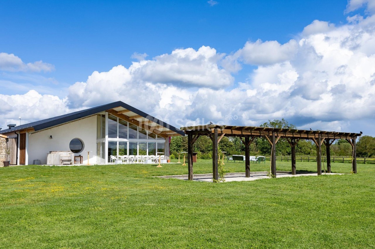 Maison moderne avec façade vitrée, pergola en bois et grand jardin verdoyant sous un ciel partiellement nuageux.