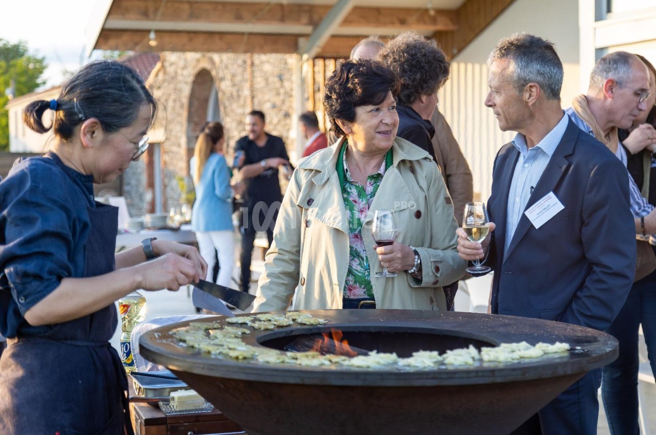 Un chef cuisine des plats sur une plancha ronde lors d'un événement en plein air avec des convives discutant.