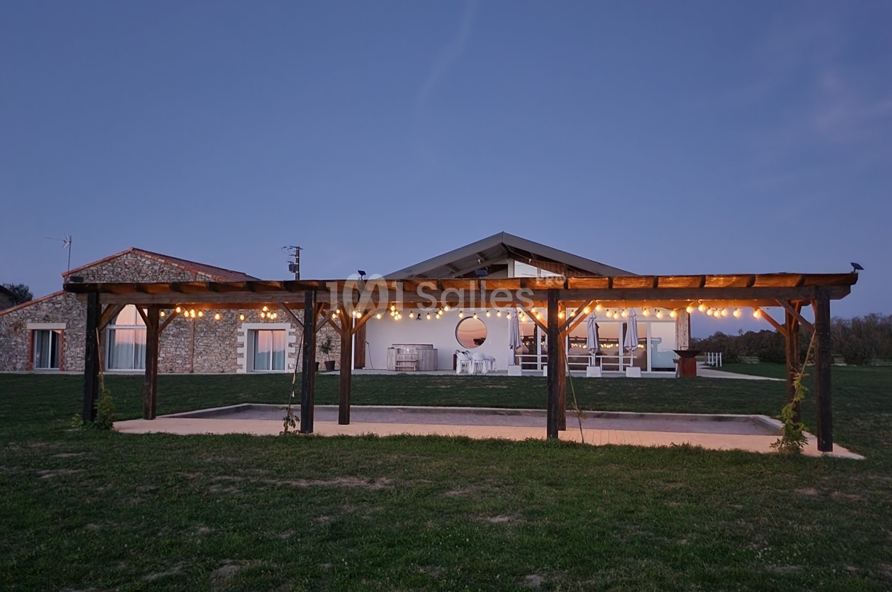 Vue d'une maison en pierre et bois avec une pergola éclairée, située dans un jardin au crépuscule.