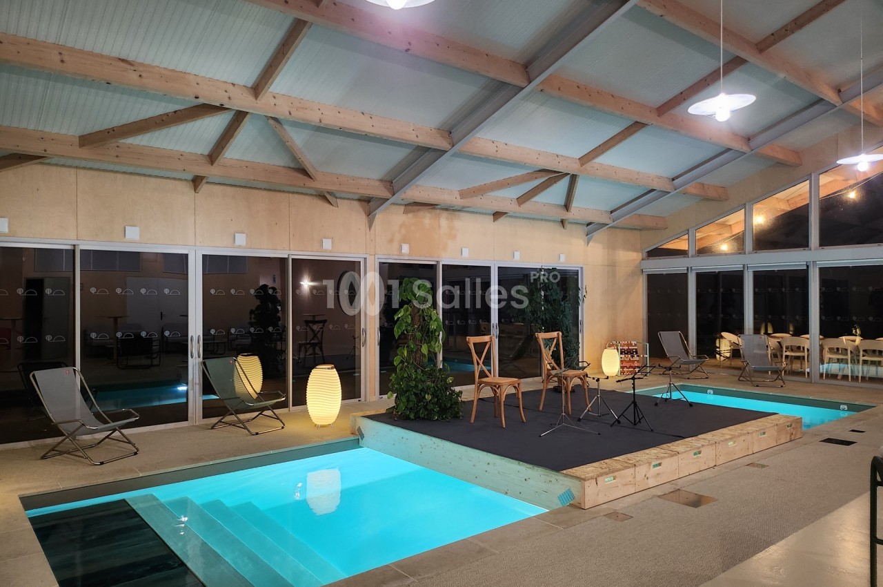 Piscine intérieure avec éclairage tamisé, entourée de chaises, plantes et d'une estrade en bois sous une structure moderne.