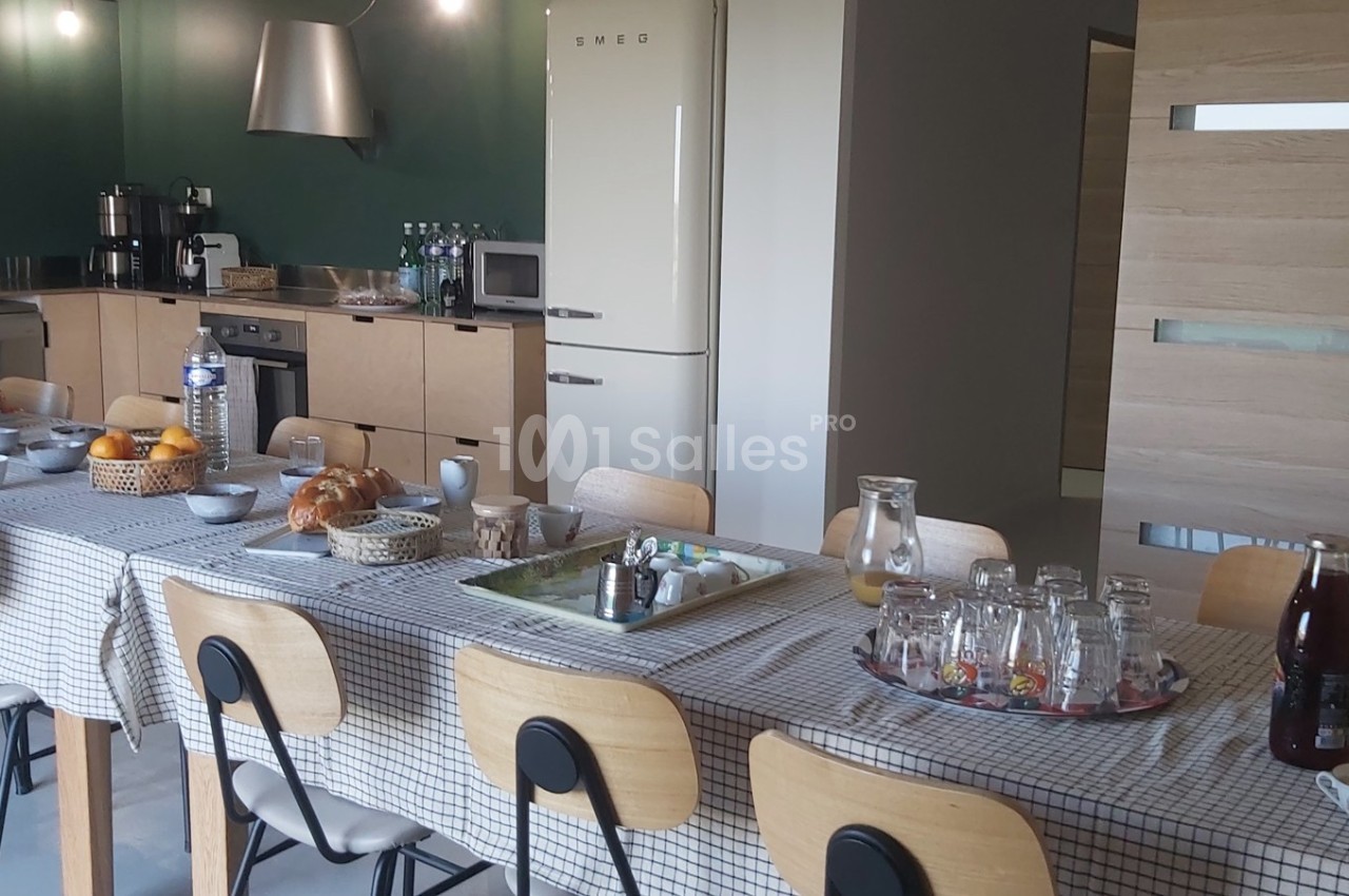 Grande table dressée avec nappes à carreaux, petits-déjeuners servis, dans une cuisine moderne avec équipements visibles.
