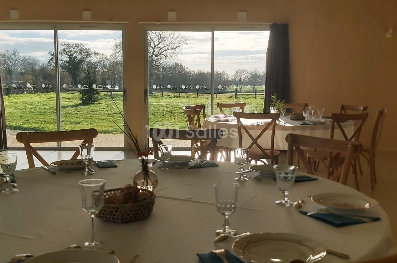 Salle lumineuse avec tables dressées, vue sur un jardin verdoyant à travers de grandes baies vitrées.