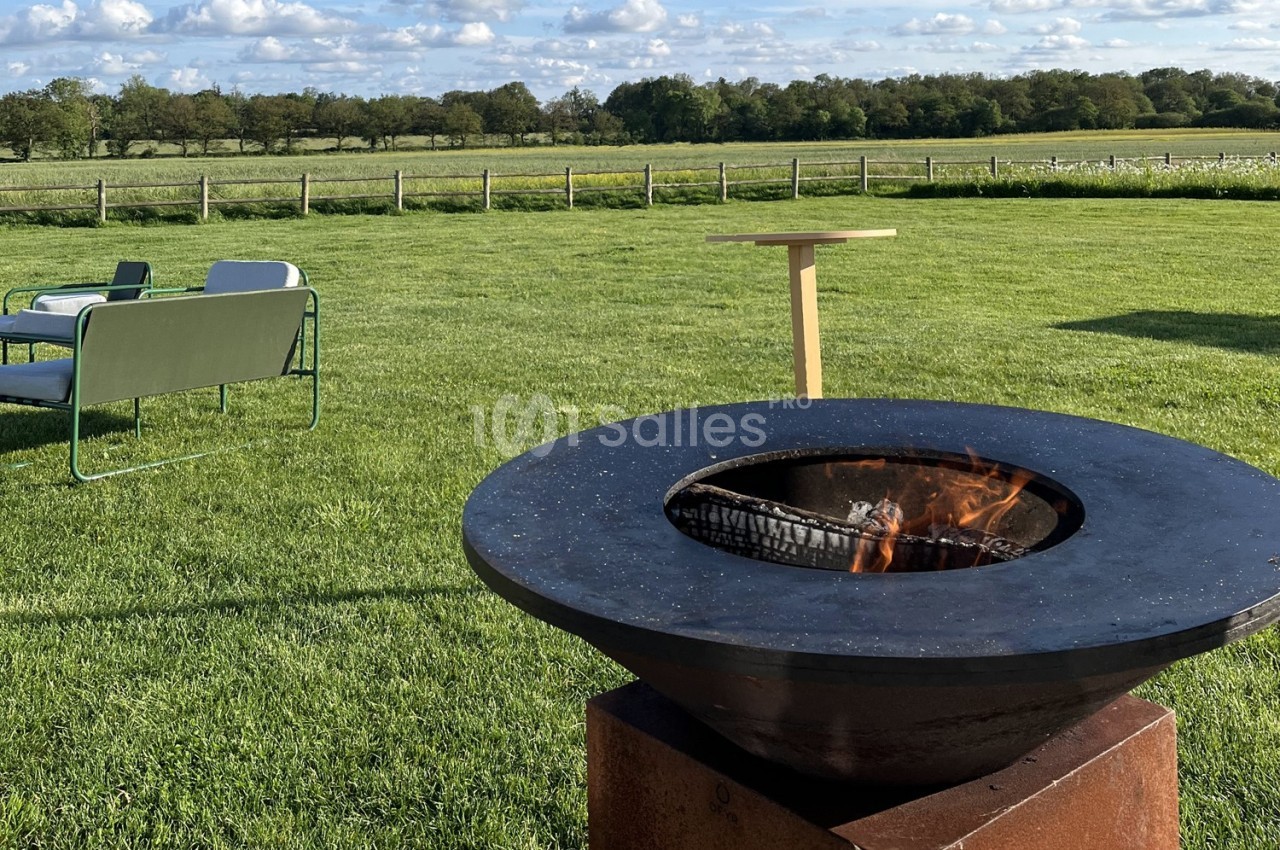 Brasero allumé au premier plan sur une pelouse, avec des sièges et une table en arrière-plan dans un cadre champêtre.
