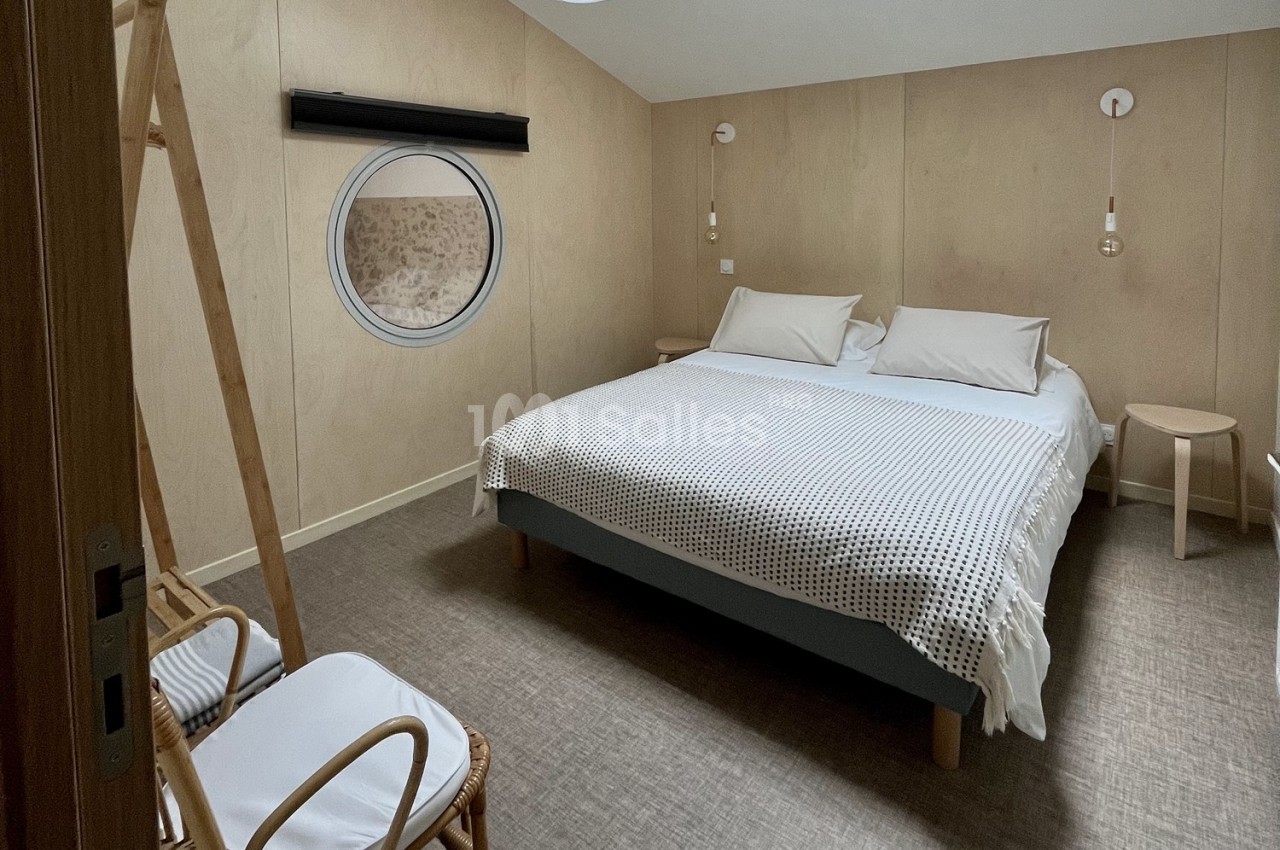 Chambre minimaliste avec lit double, murs en bois clair, hublot rond et mobilier en bois naturel.