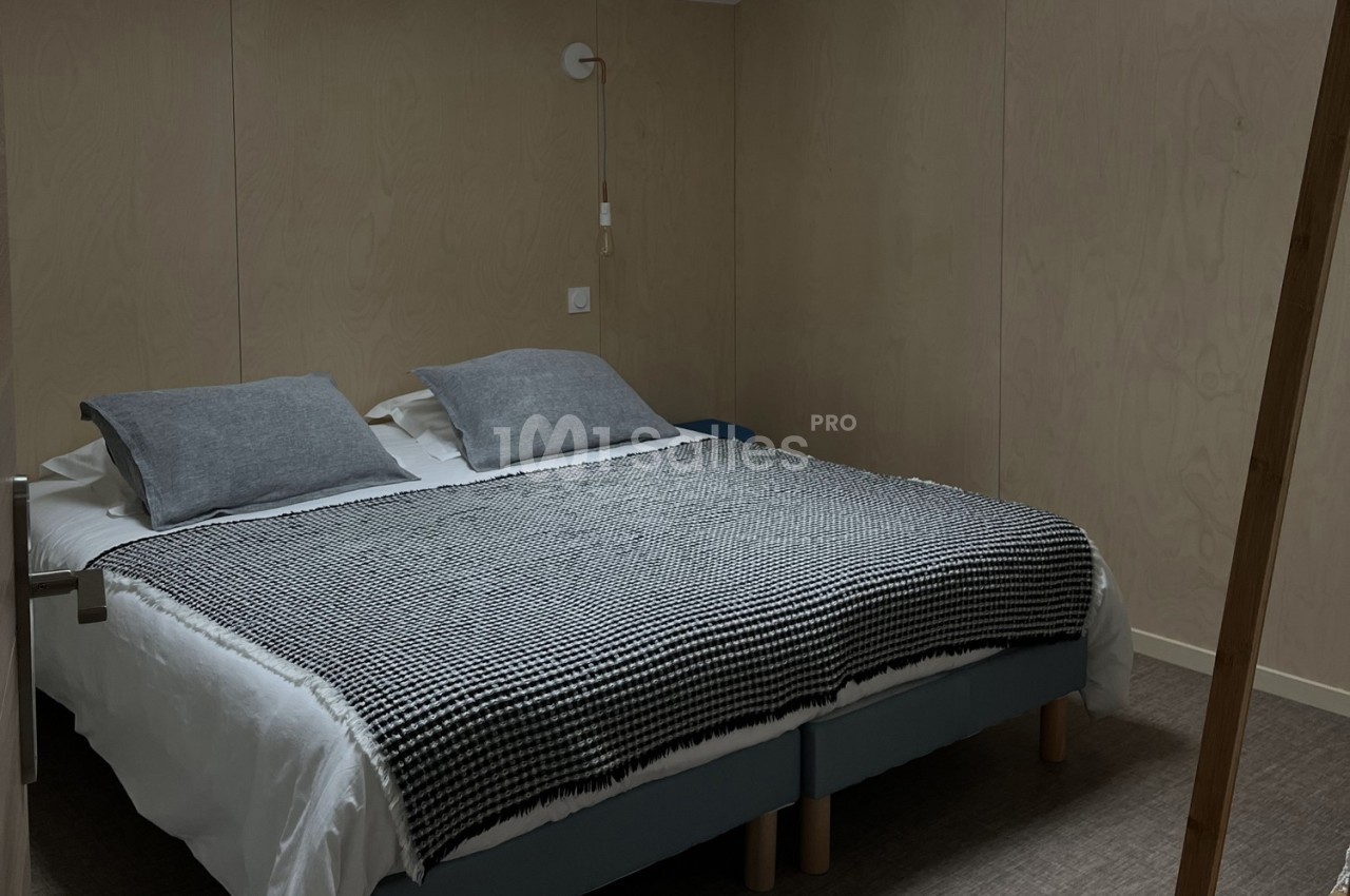 Lit double avec draps blancs, plaid à motifs noirs et blancs, et coussins gris dans une chambre aux murs en bois clair.