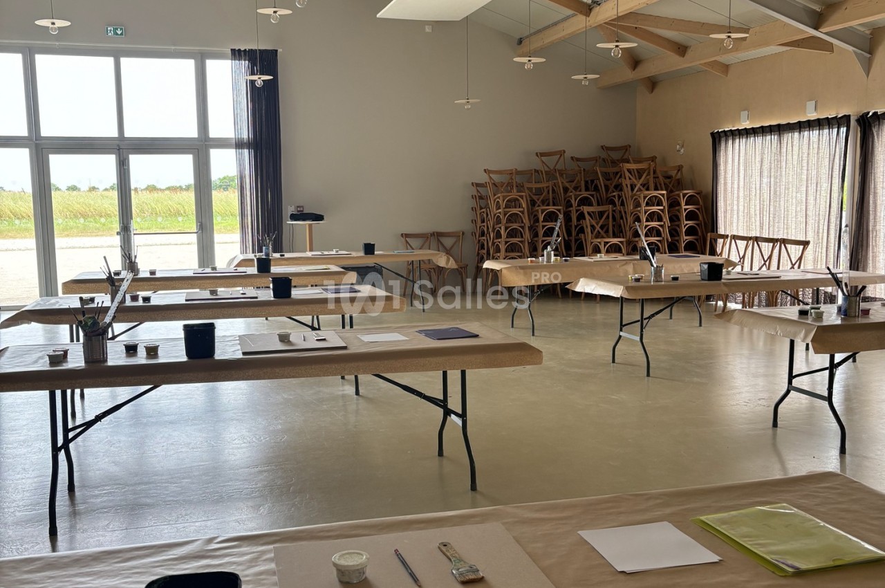 Salle lumineuse avec tables alignées, couvertes de matériel d'art, et des chaises empilées dans un coin.