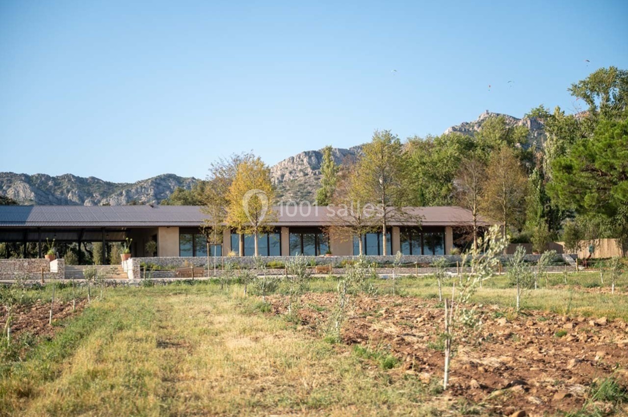 Bâtiment moderne entouré de champs cultivés, avec des montagnes et des arbres en arrière-plan sous un ciel dégagé.