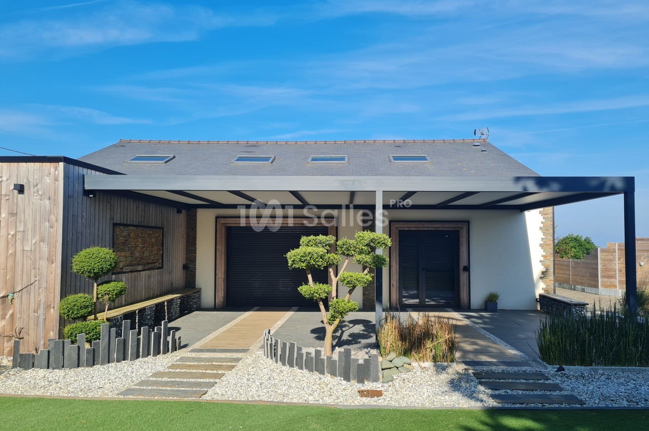 Maison moderne avec toit en ardoise, pergola métallique, jardin aménagé et allée en gravier sous un ciel bleu.