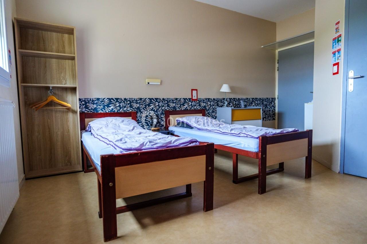 Deux lits simples avec draps dans une chambre sobre, équipée d'une armoire ouverte et d'une table de chevet.