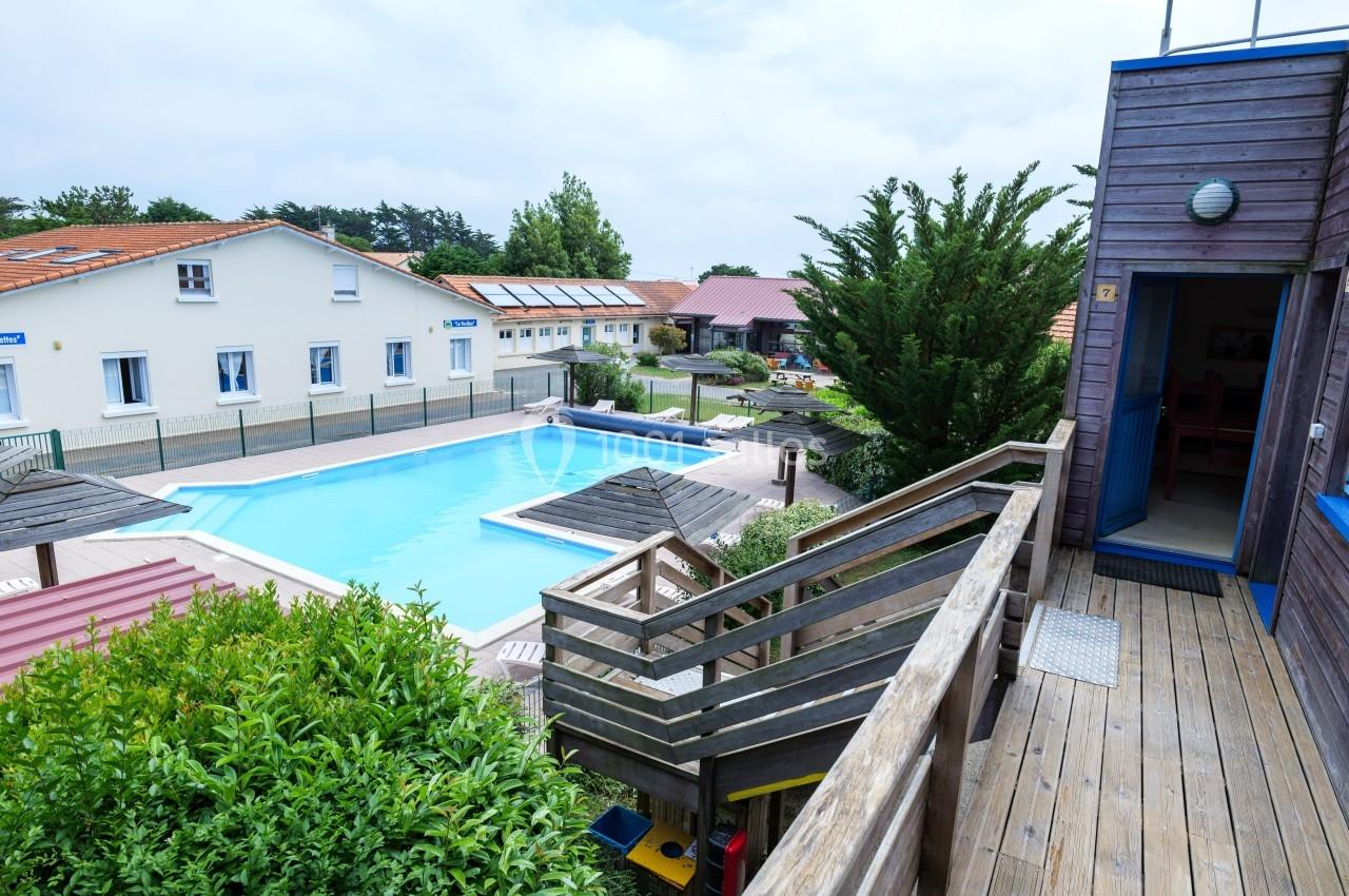 Vue d'une piscine extérieure entourée de bâtiments, avec des parasols en bois et un escalier en premier plan.