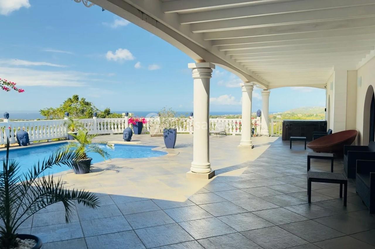 Terrasse couverte avec piscine, vue sur la mer et végétation, entourée de colonnes blanches et mobilier extérieur.