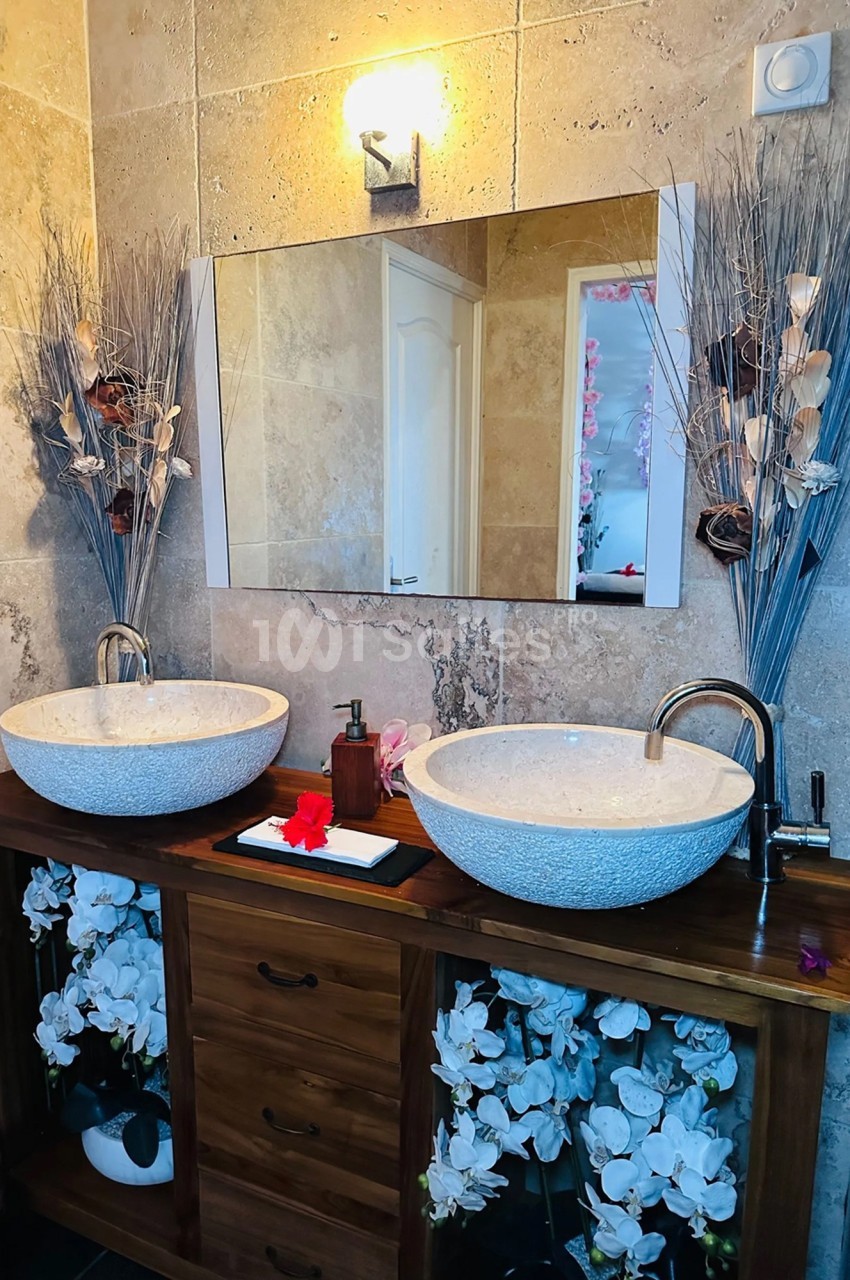Double vasque en pierre sur meuble en bois, décorée de fleurs blanches et sèches, avec miroir et éclairage mural.