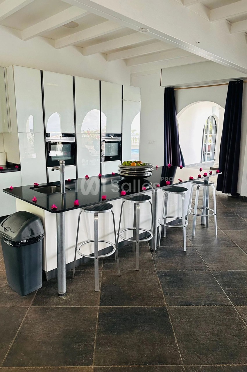 Cuisine moderne avec îlot central noir, tabourets métalliques, décorée de petites fleurs roses et éclairée par des fenêtres…
