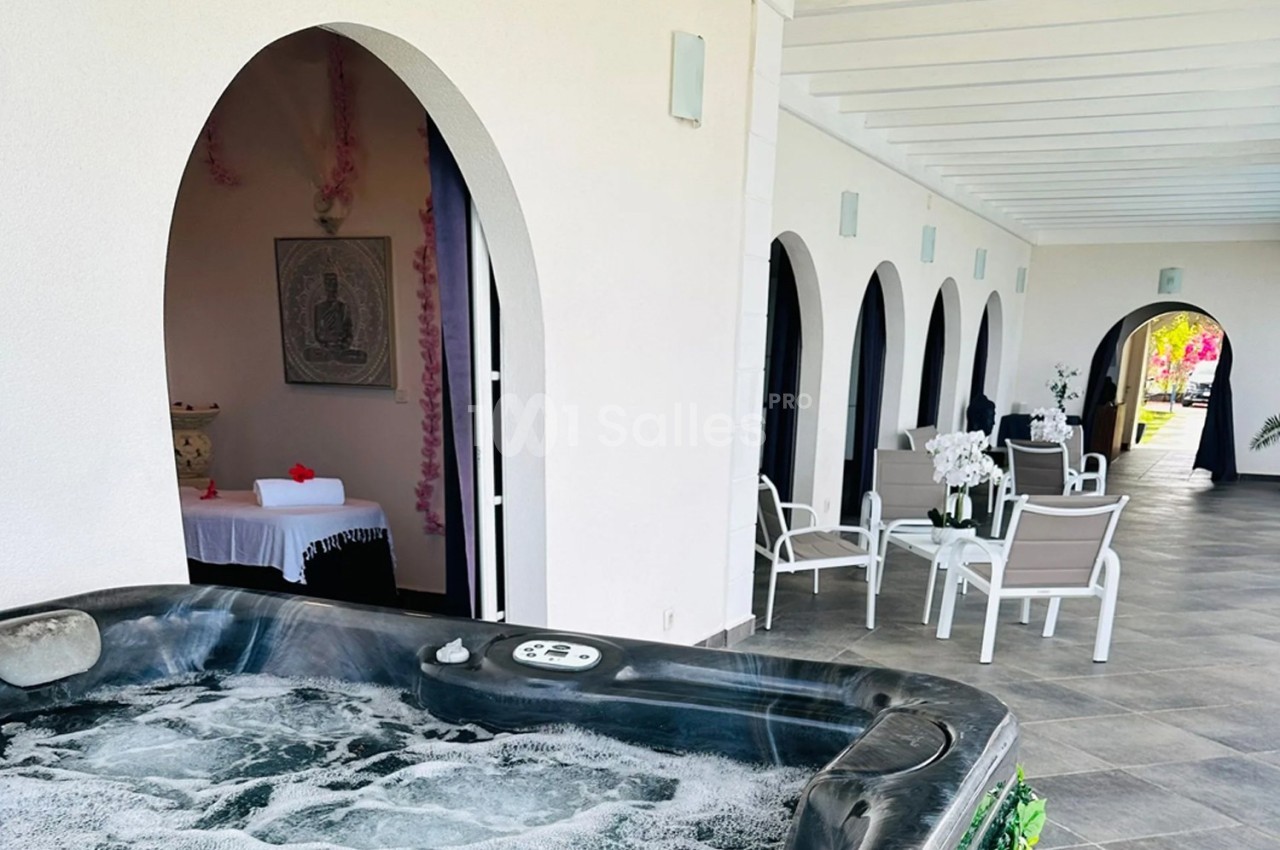 Bain à remous en premier plan avec terrasse couverte, chaises blanches et espace de massage en arrière-plan.
