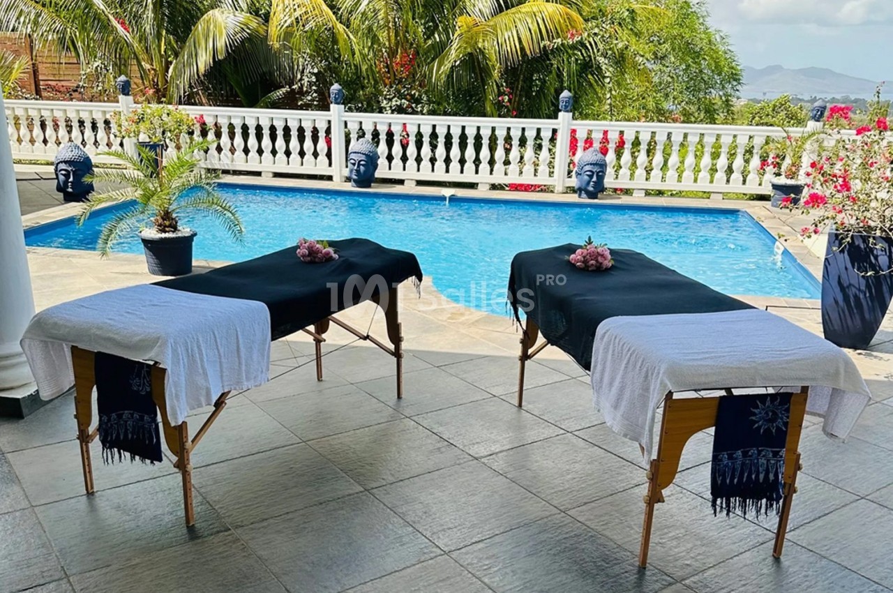 Deux tables de massage couvertes de draps noirs et blancs, placées près d'une piscine entourée de végétation tropicale.