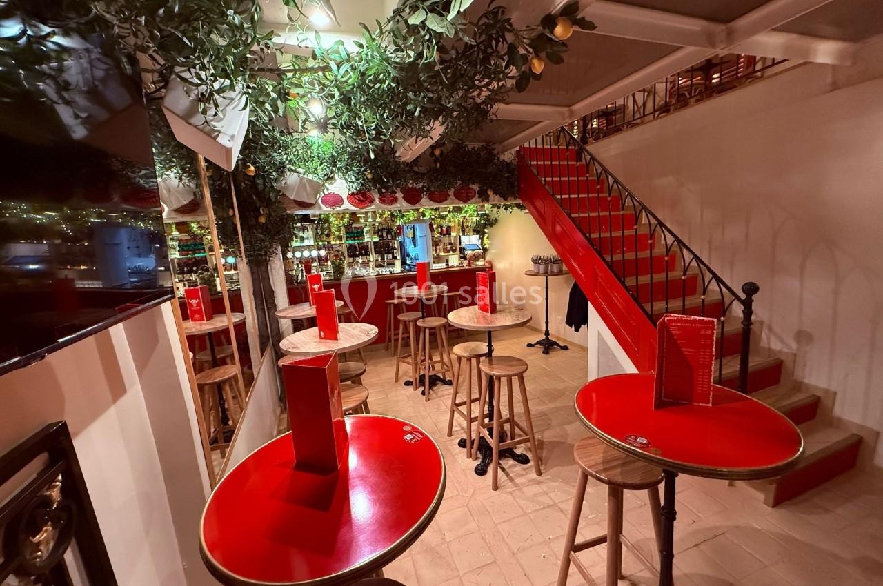 Salle de bar avec tables rouges, tabourets en bois, escalier rouge et décoration végétale suspendue.