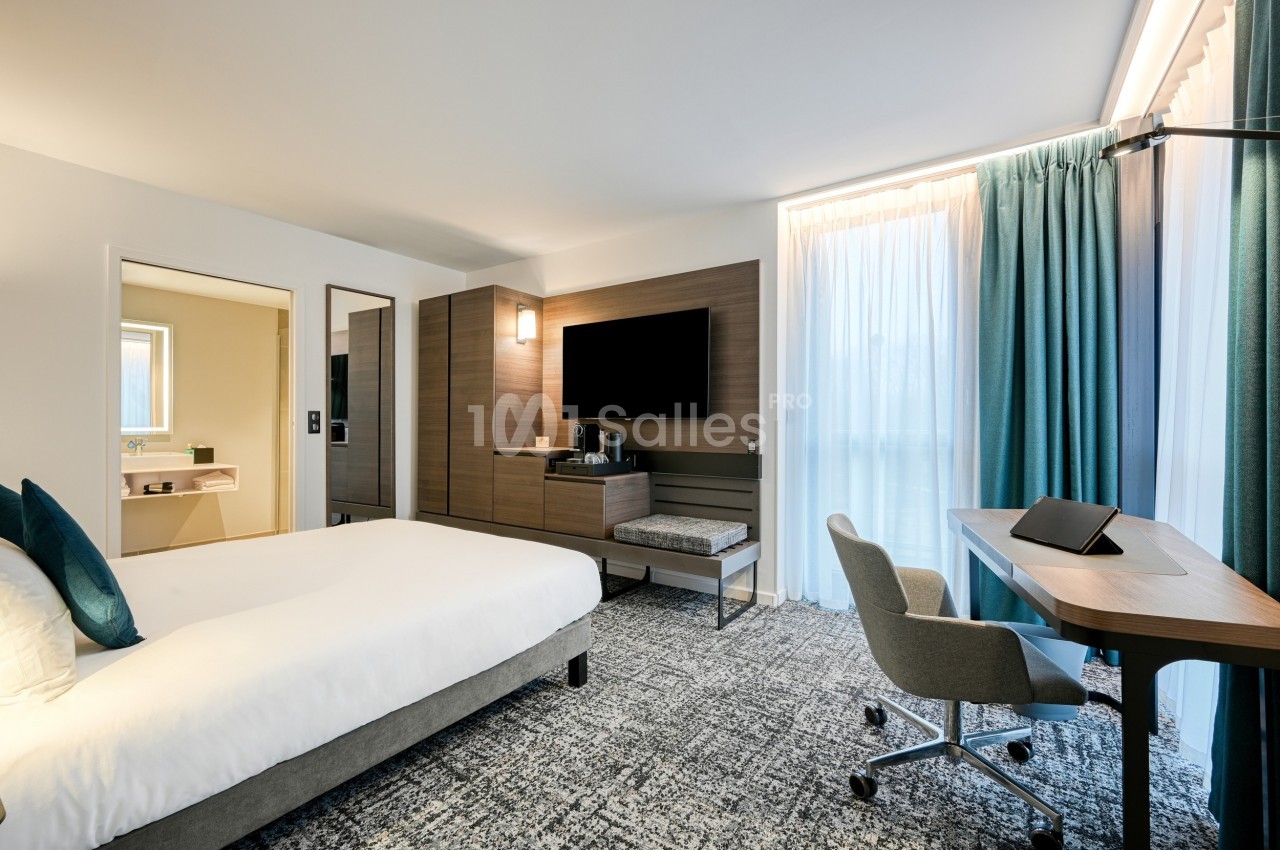 Chambre d'hôtel moderne avec lit double, bureau, télévision murale, salle de bain attenante et grandes fenêtres avec rideaux.
