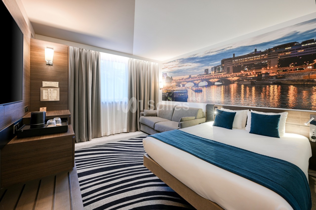 Chambre d'hôtel moderne avec lit double, canapé, bureau, éclairage doux et grande photo murale d'un pont illuminé.