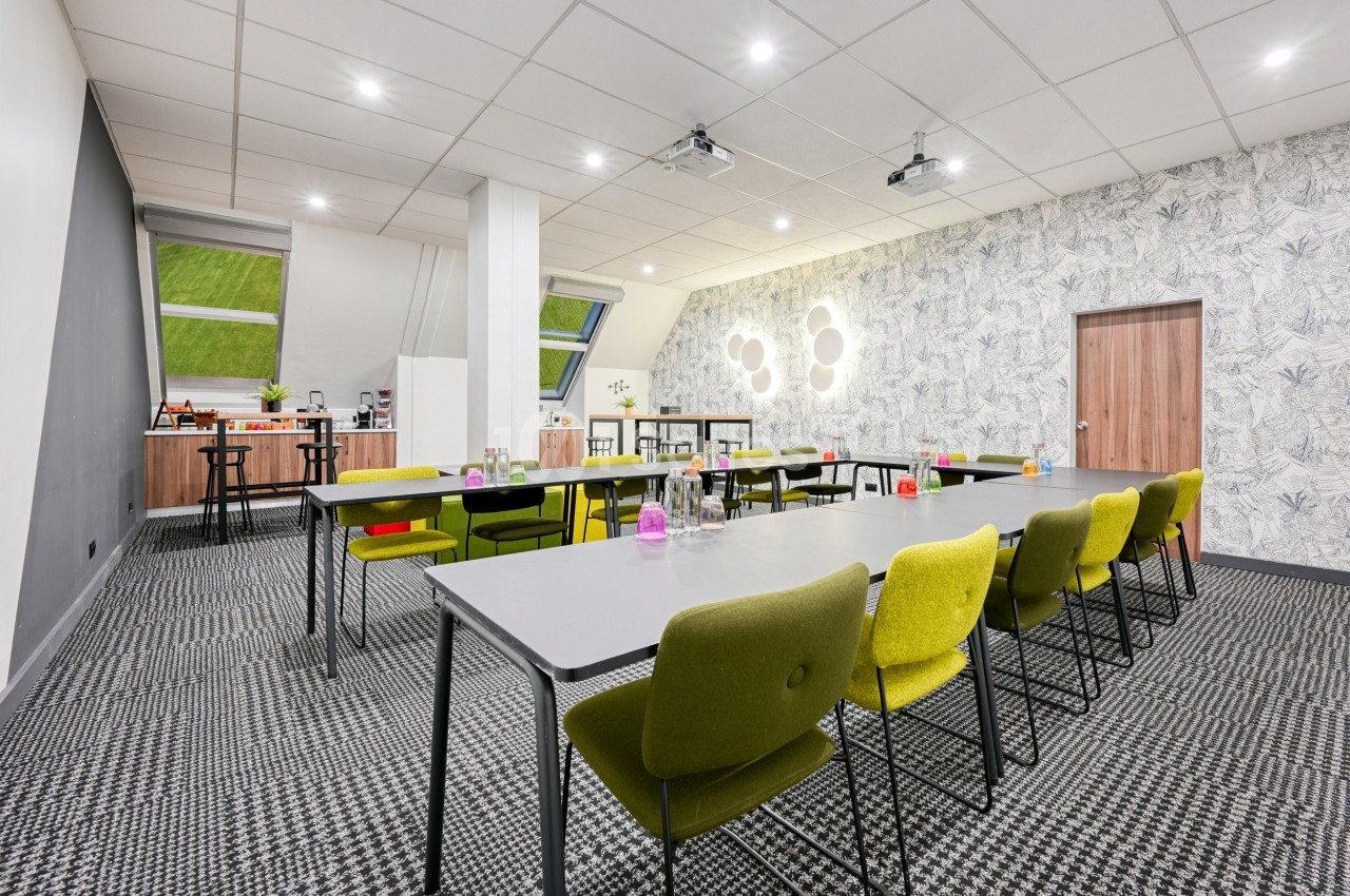 Salle de réunion lumineuse avec tables grises, chaises vertes, projecteurs au plafond et coin café en arrière-plan.