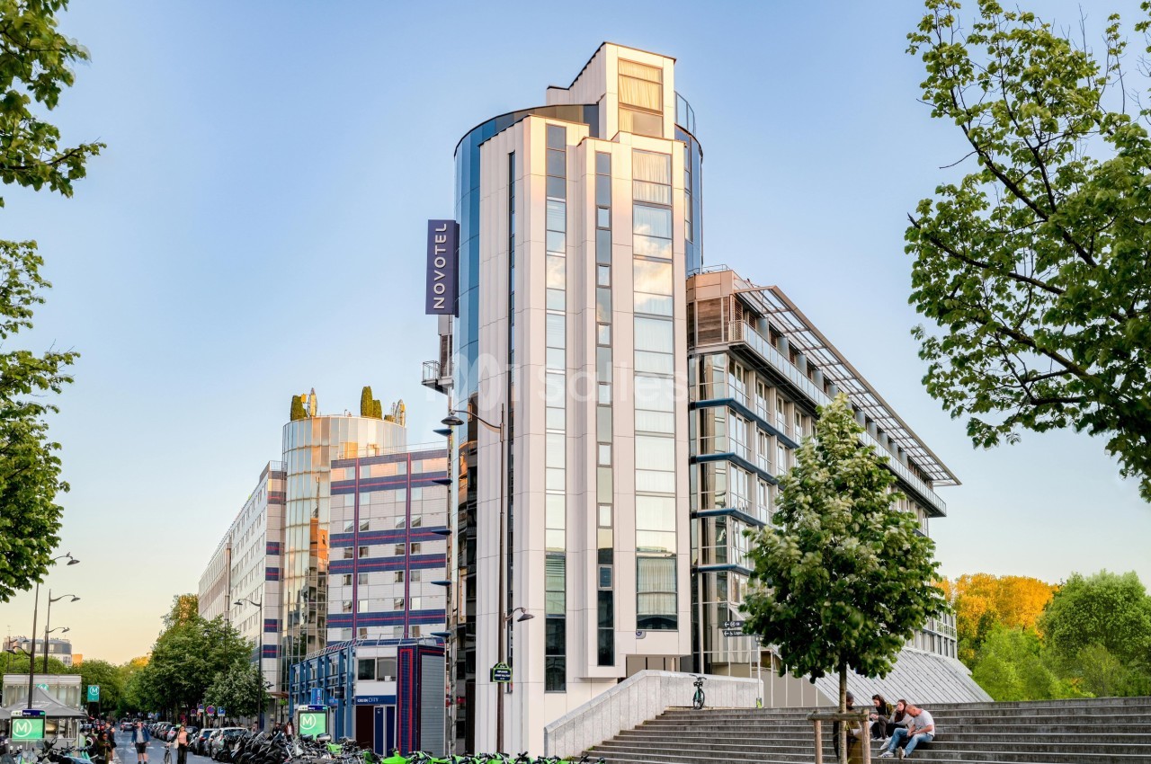 Bâtiment moderne en verre et métal avec un panneau ’Novotel’, entouré d'arbres et de vélos stationnés.