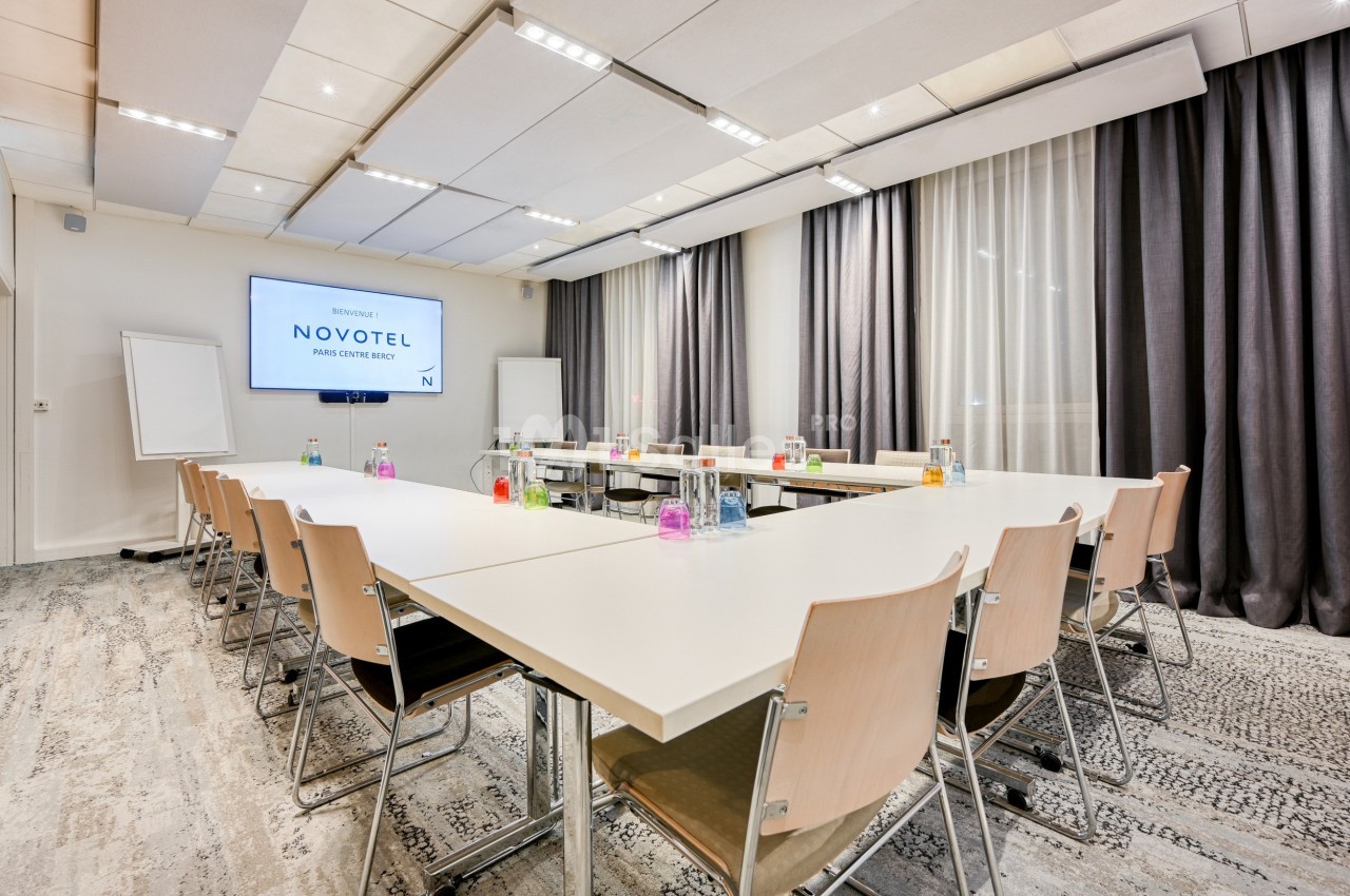Salle de réunion lumineuse avec tables en configuration rectangulaire, chaises en bois clair et écran de présentation.