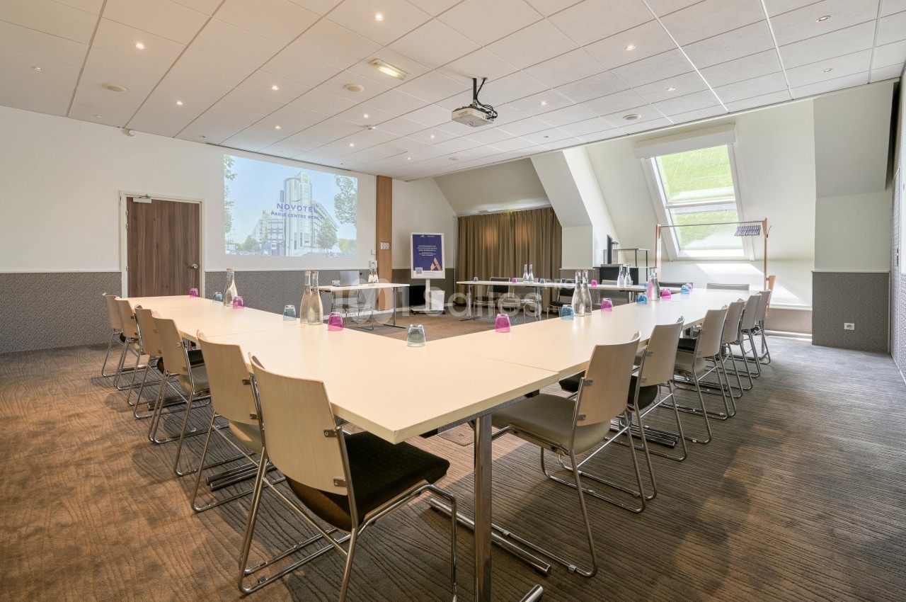 Salle de réunion lumineuse avec tables disposées en U, chaises en bois, projecteur et écran de présentation.