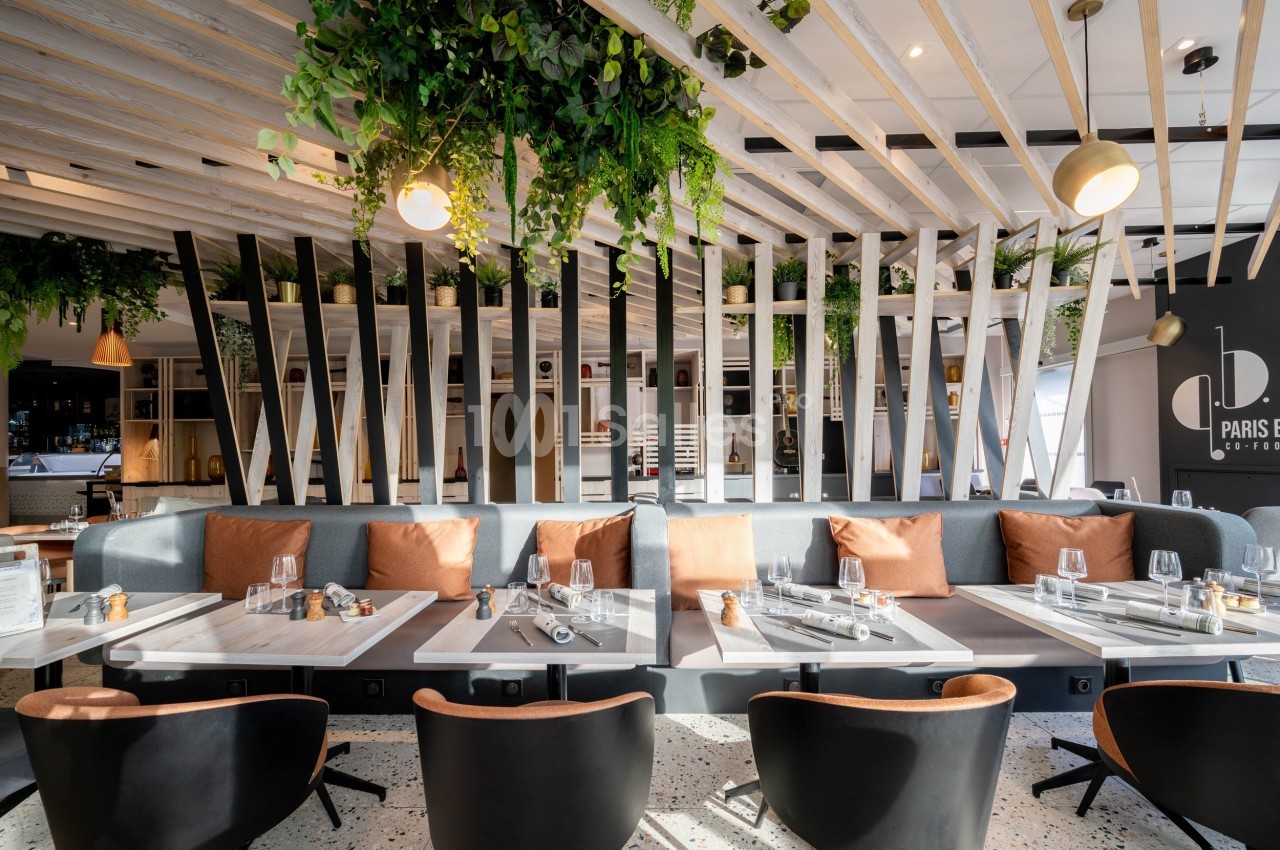 Salle de restaurant moderne avec tables dressées, banquettes grises, coussins orange et plantes suspendues.