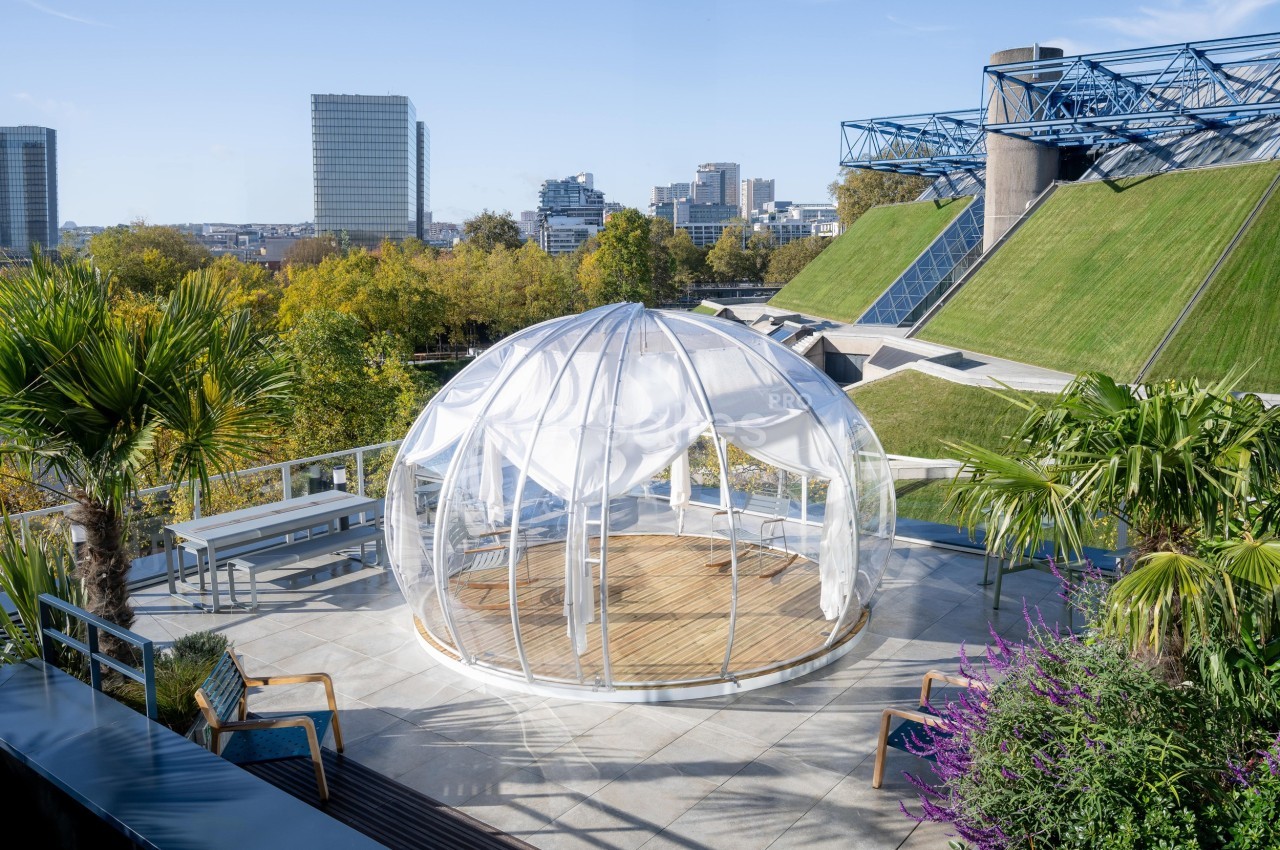 Dôme transparent installé sur une terrasse entourée de végétation, avec vue sur des bâtiments urbains modernes.