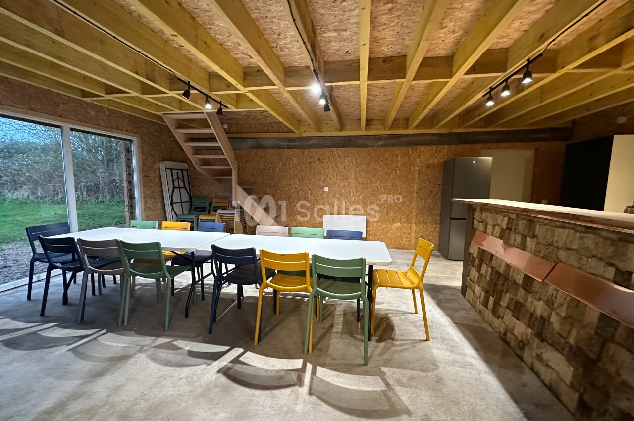 Salle avec une grande table entourée de chaises colorées, un escalier en bois et un bar en briques dans un espace lumineux.
