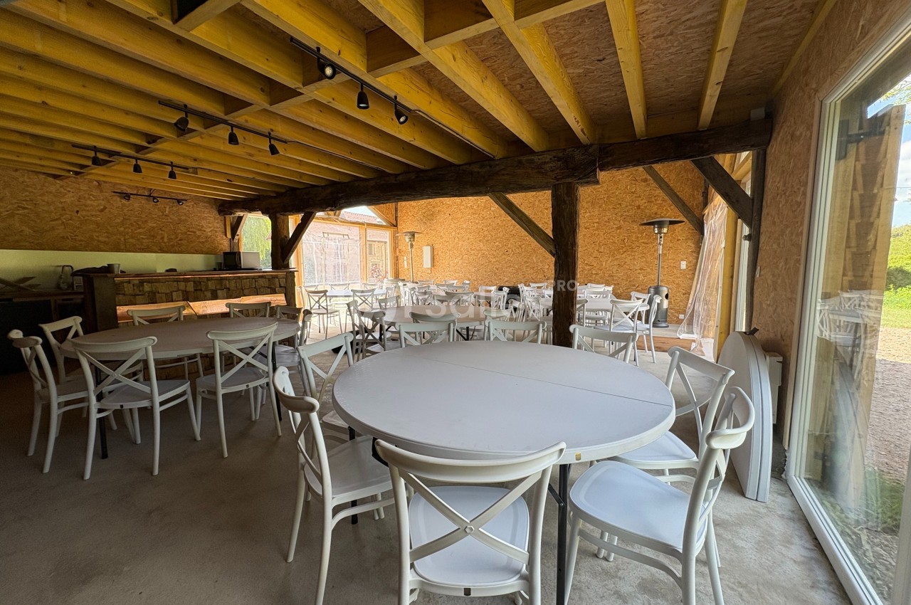 Salle avec des tables rondes et chaises blanches, charpente en bois apparente et vue sur une cour extérieure.