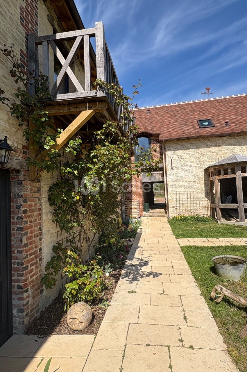 Cour pavée bordée de bâtiments en pierre avec balcon en bois, jardin verdoyant et ciel bleu en arrière-plan.