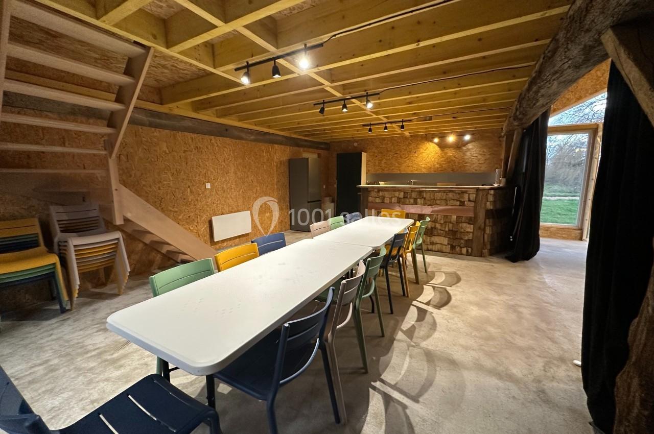 Salle avec une grande table entourée de chaises colorées, éclairée par des spots, avec un escalier et un bar en arrière-plan.