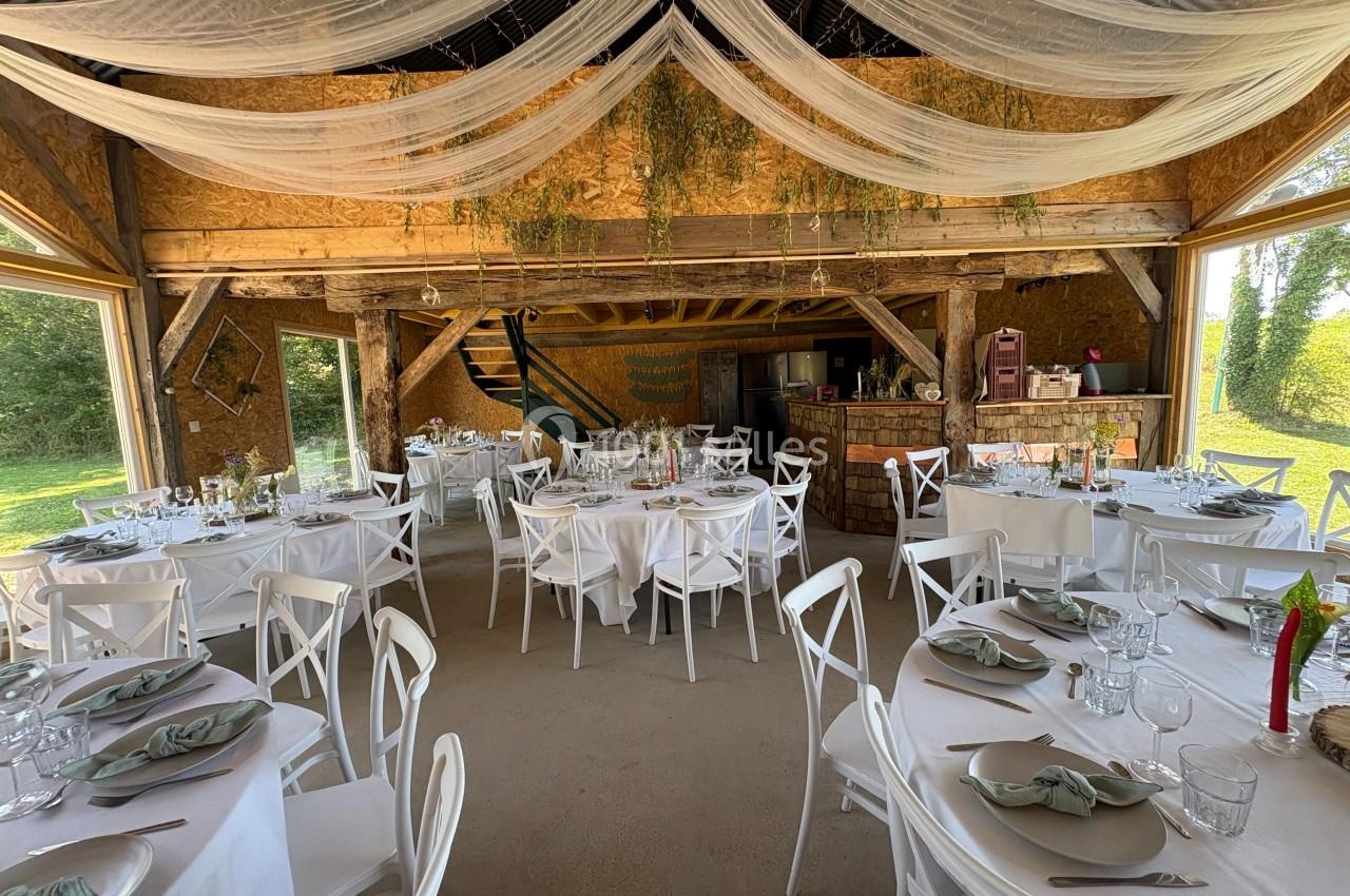 Salle de réception rustique avec tables rondes dressées, chaises blanches et décorations suspendues en tissu et végétation.