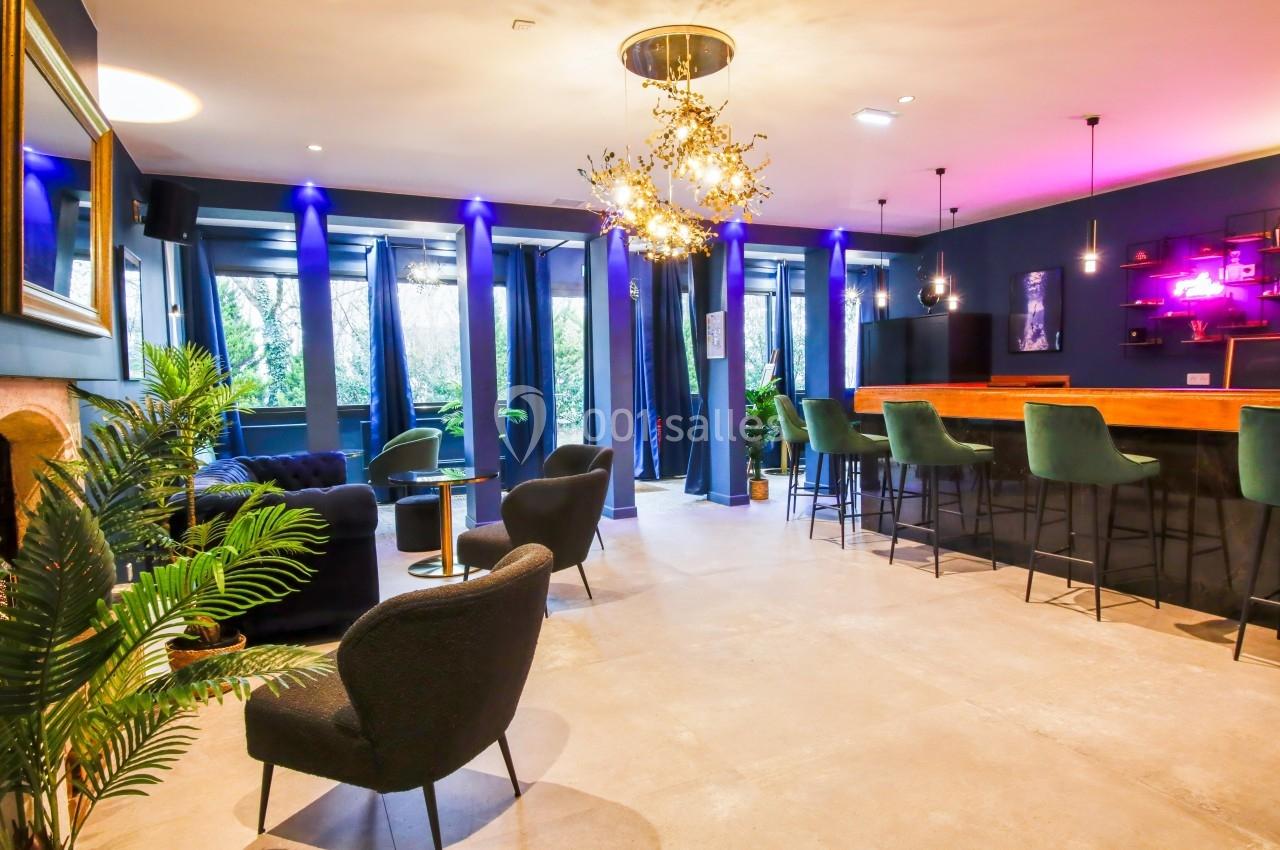 Salle lumineuse avec bar en bois, fauteuils noirs, plantes, et éclairage violet créant une ambiance moderne et chaleureuse.