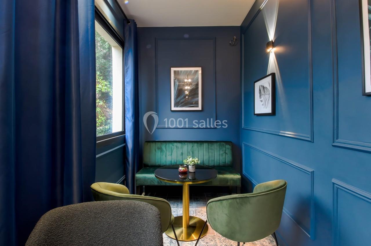 Coin salon élégant avec murs bleus, canapé vert, deux fauteuils assortis et une table ronde dorée près d'une fenêtre.