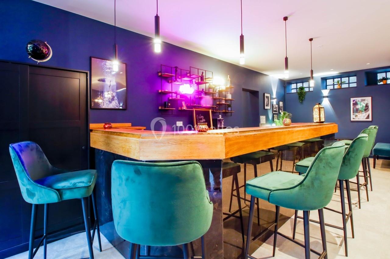 Bar moderne avec comptoir en bois, tabourets verts, éclairage coloré et décorations murales dans une ambiance chaleureuse.