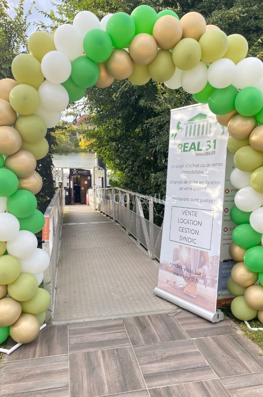 Arche décorative en ballons verts, blancs et dorés devant une passerelle menant à un bâtiment, avec une bannière d'agence…