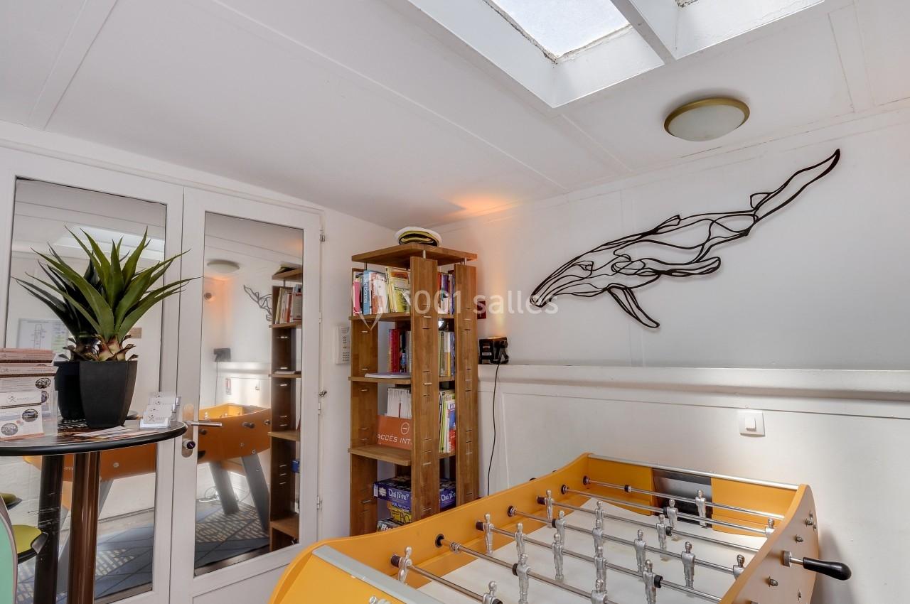 Salle lumineuse avec baby-foot jaune, bibliothèque en bois, plante en pot et décoration murale en forme de baleine.