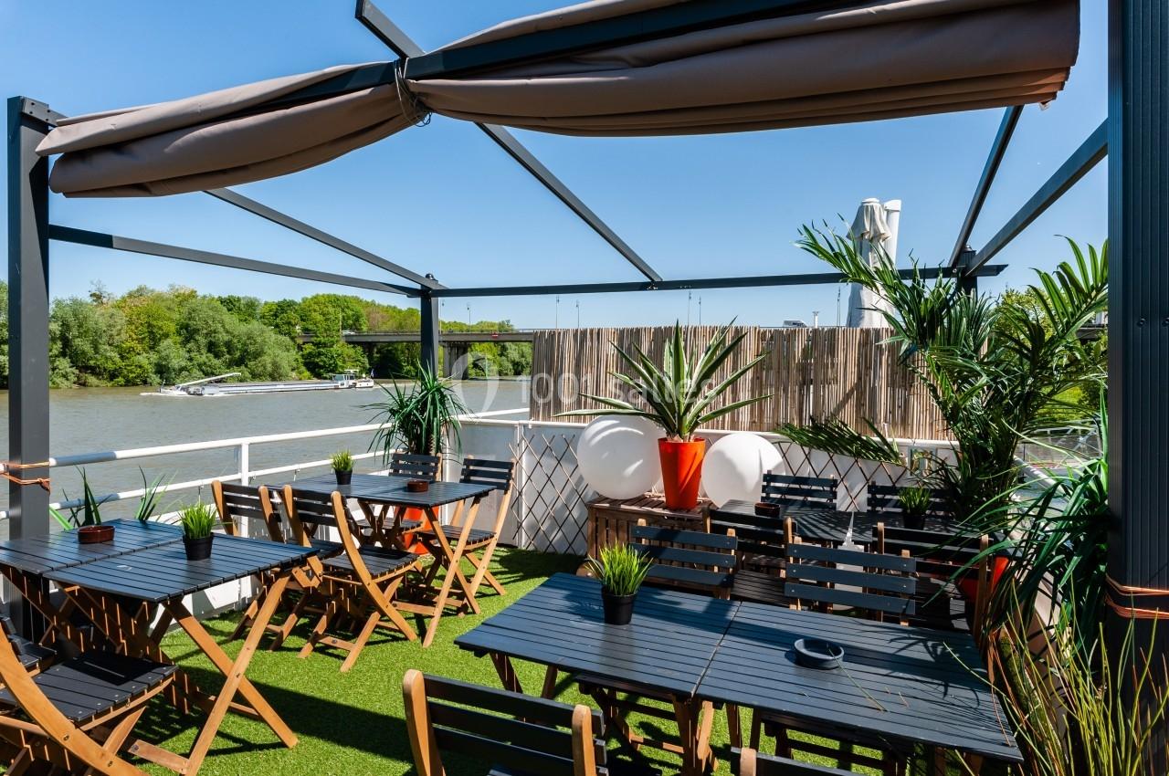 Terrasse extérieure avec tables, chaises et plantes, offrant une vue sur une rivière sous un ciel dégagé.