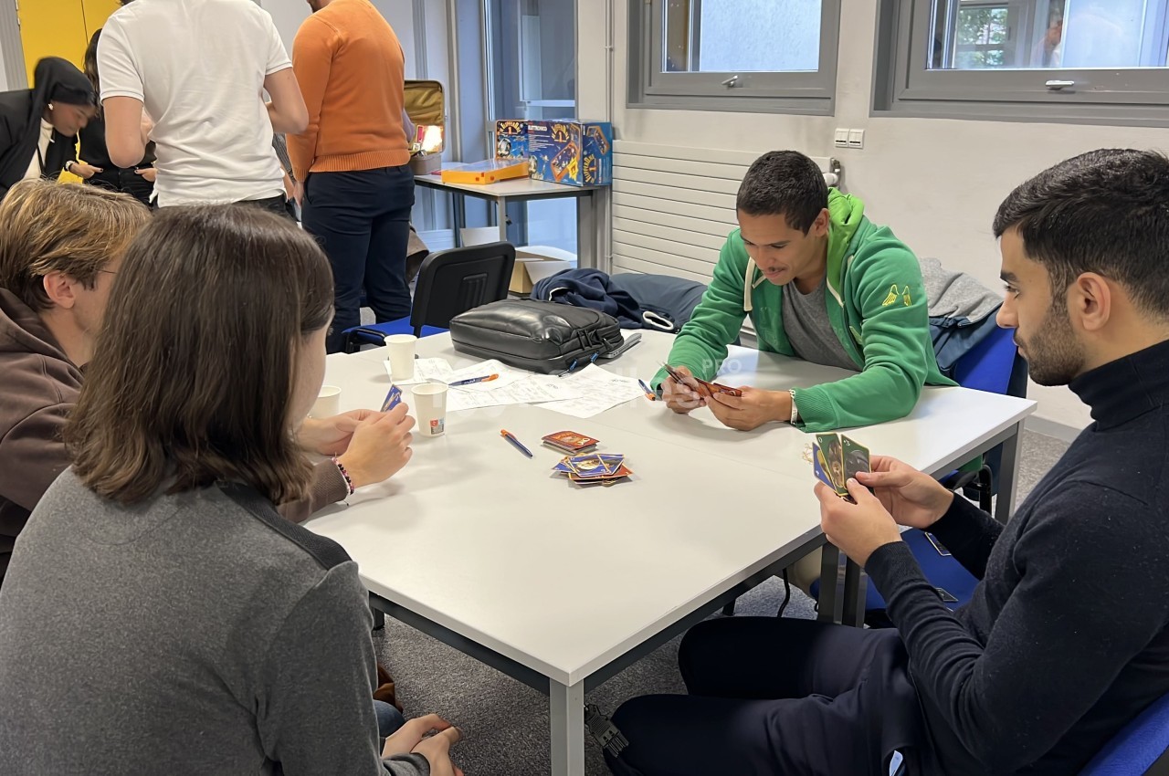 Quatre personnes jouent à un jeu de cartes autour d'une table dans une salle lumineuse avec d'autres personnes en arrière…