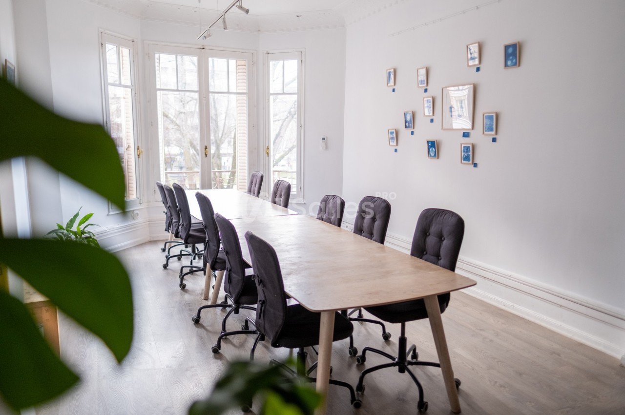 Salle de réunion lumineuse avec une grande table en bois, des chaises noires et des cadres accrochés au mur.