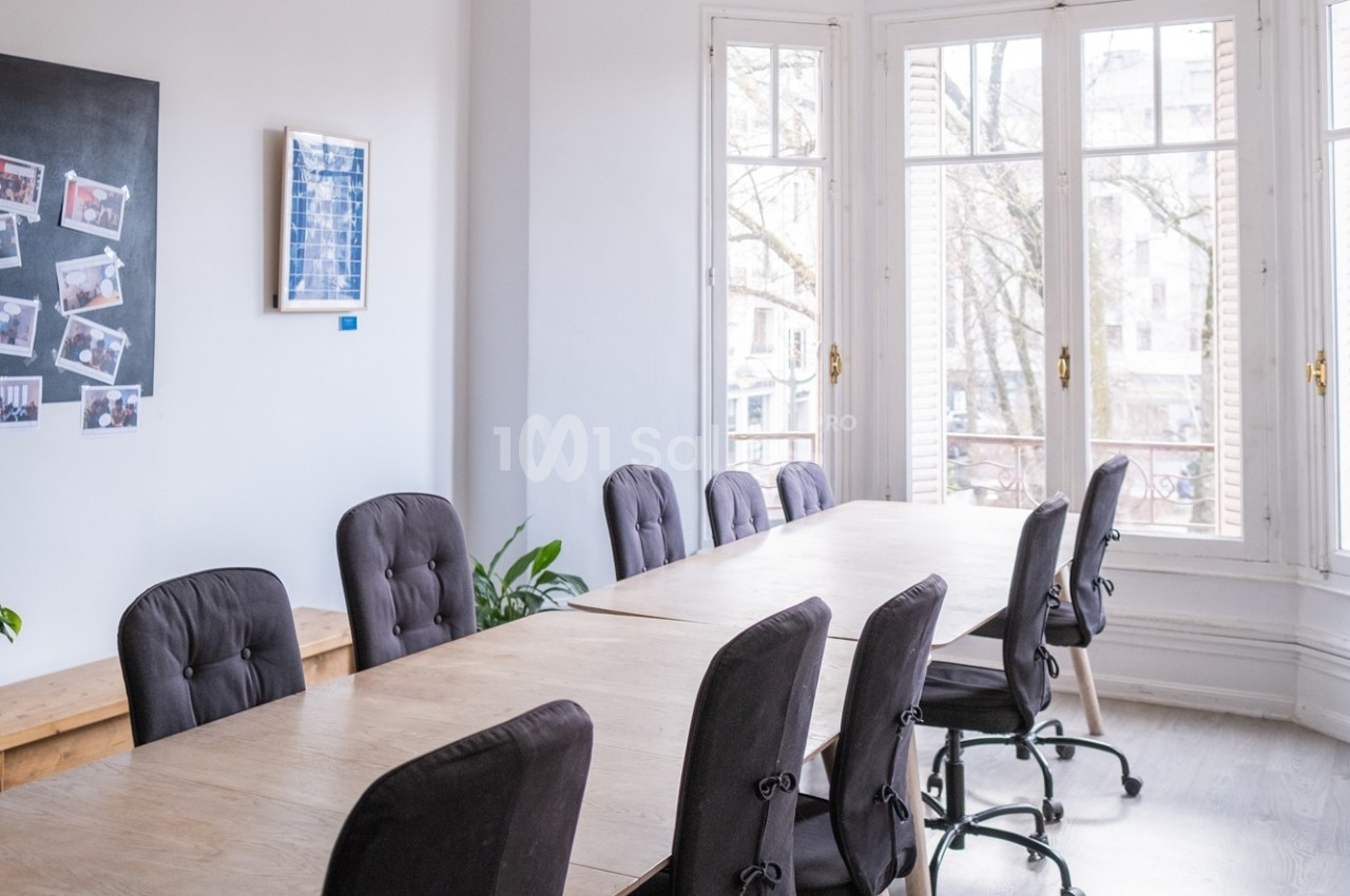 Salle de réunion lumineuse avec une grande table en bois entourée de chaises, près de fenêtres donnant sur l'extérieur.