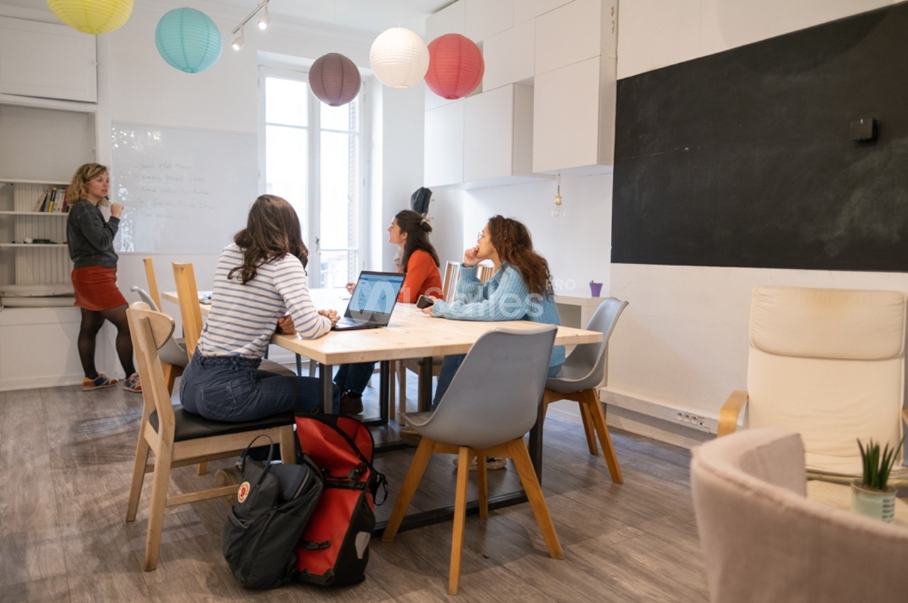 Quatre personnes discutent autour d'une table dans une salle lumineuse, avec une femme écrivant au tableau blanc.