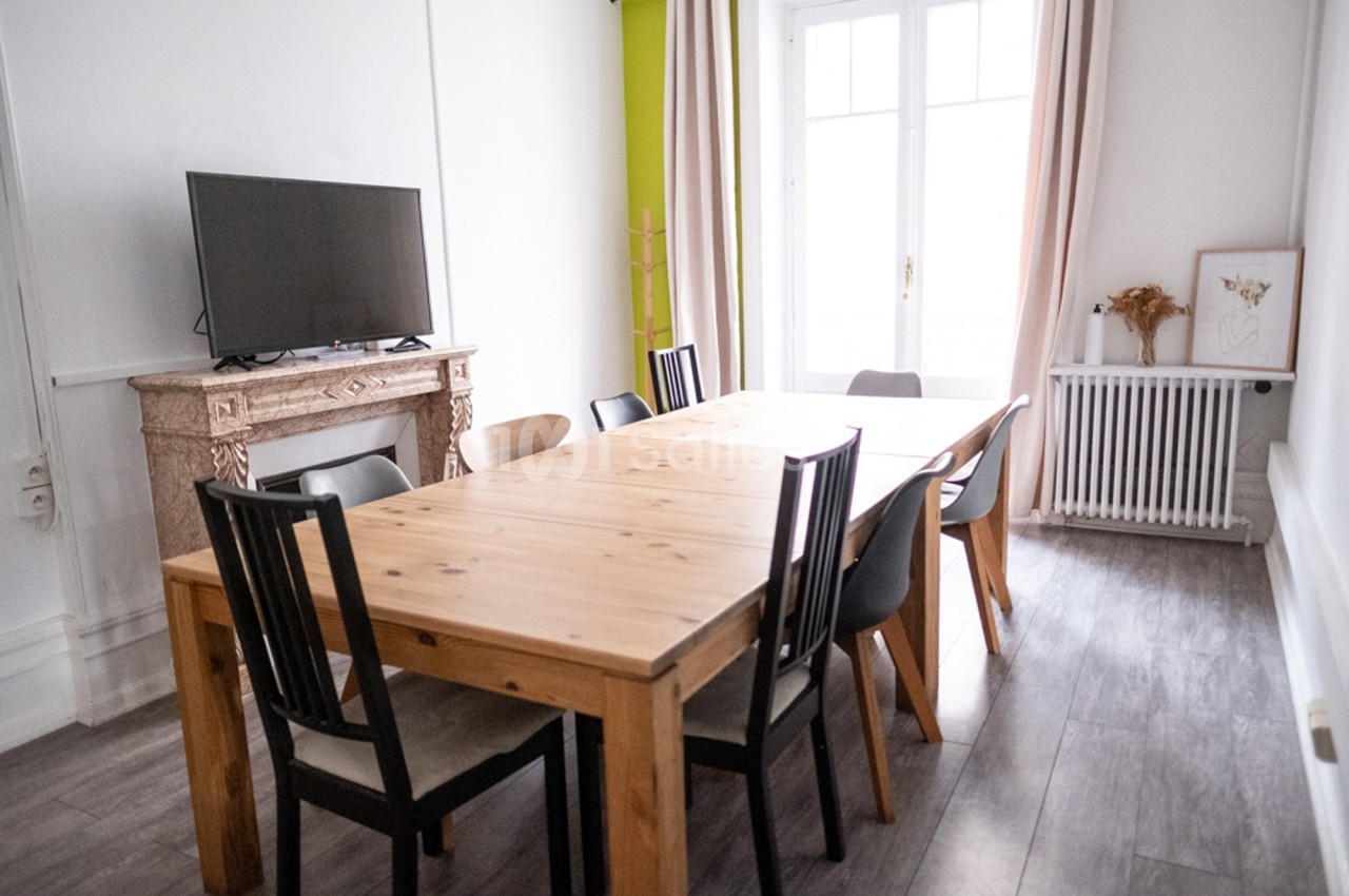 Salle à manger lumineuse avec table en bois, chaises noires et grises, cheminée, télévision et grande fenêtre.