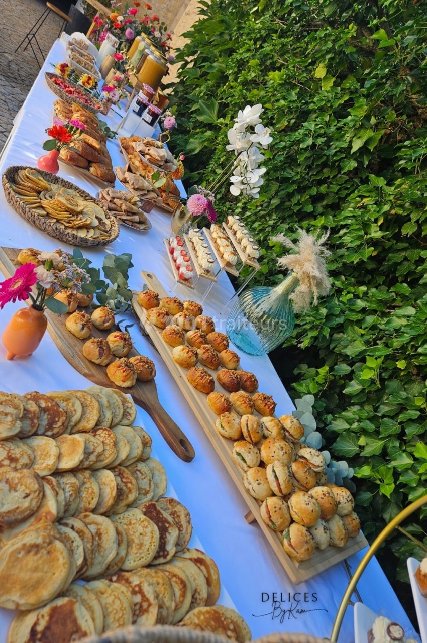 Table garnie de pâtisseries variées, crêpes, muffins et décorations florales, disposée en extérieur près d'un mur végétal.
