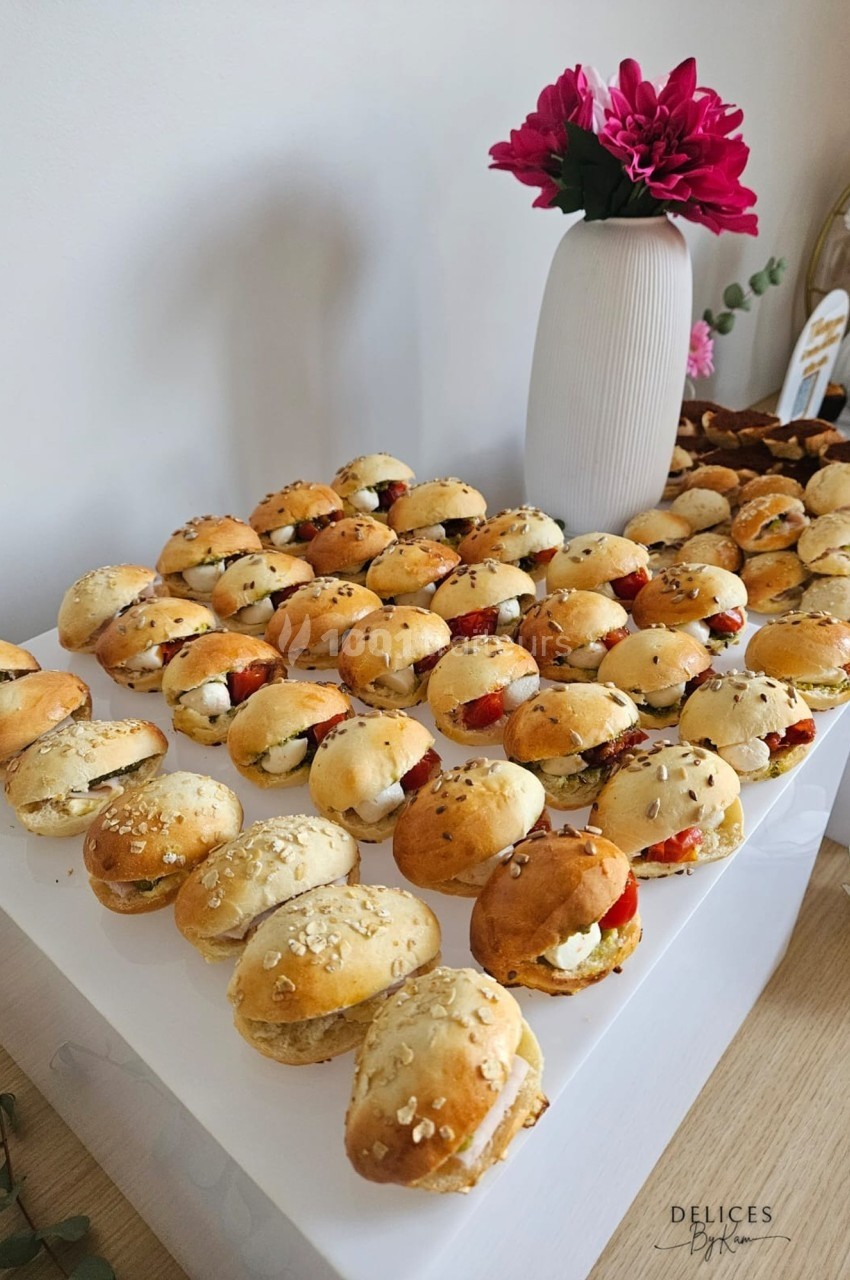 Plateau de mini-sandwichs variés disposés sur une table, avec un vase de fleurs roses en arrière-plan.