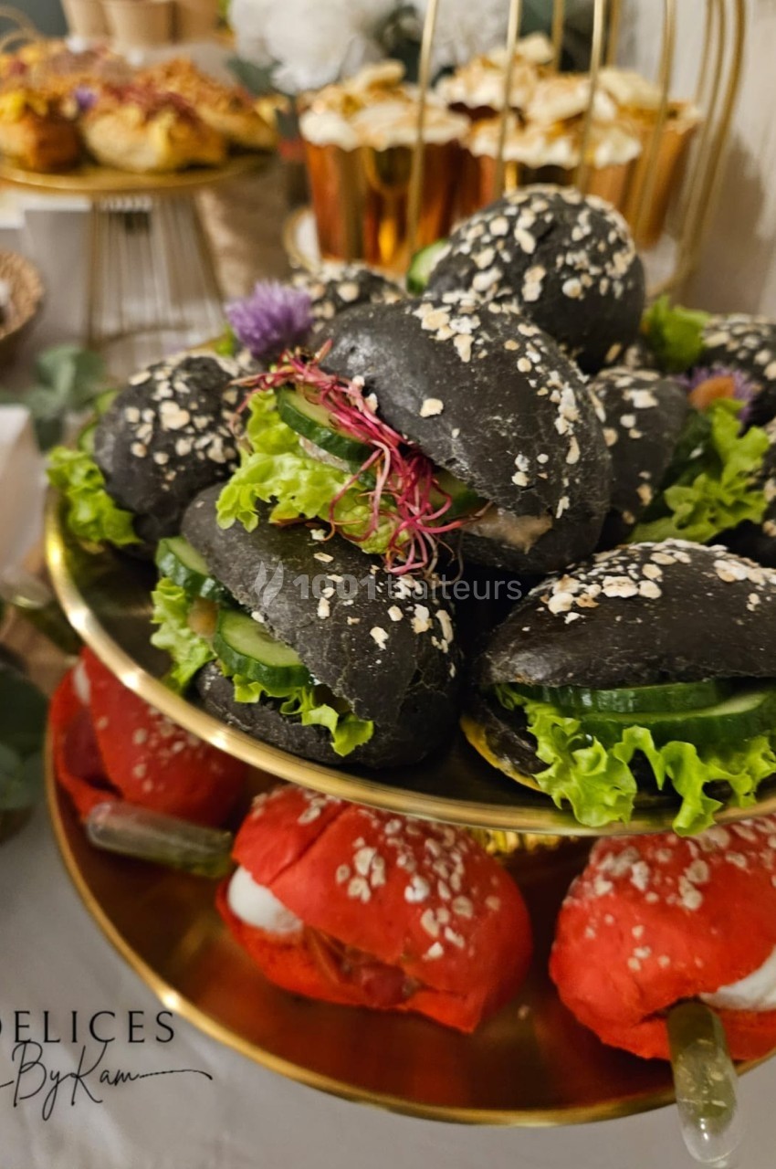 Plateau de mini-burgers noirs garnis de salade, concombre et pousses, avec des pains rouges en arrière-plan.
